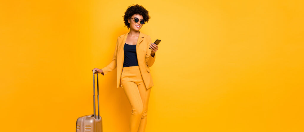 woman in yellow outfit in front of yellow background holding a mobile phone in one hand and a suitcase in the other